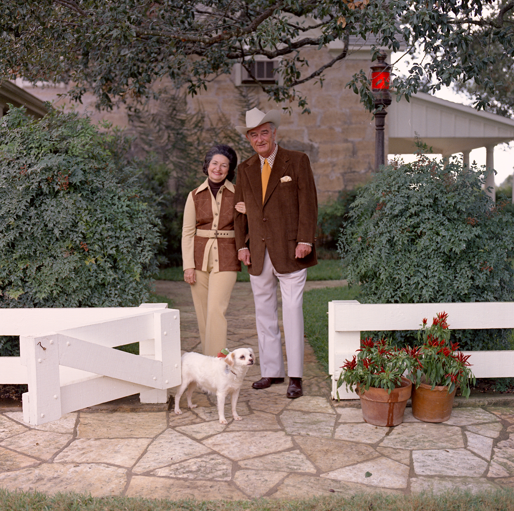 Lady Bird and Lyndon Johnson in Western wear with a small white dog, Yuki, standing by a low, white wooden fence in front of a ranch house.